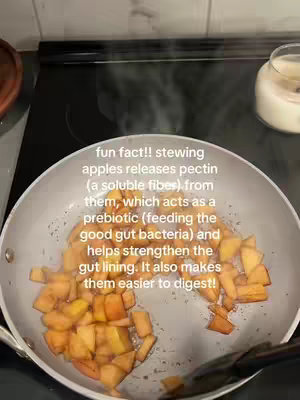 A top-down shot shows a stainless steel pan on a stovetop. Inside the pan, a small quantity of stewed apples is visible. The apples are golden-brown and glistening, indicating they are cooked. Steam rises from the pan, suggesting the apples are still warm. Partially visible in the background are parts of the stovetop surface and a jar with a white substance, possibly a candle. The overall impression is one of preparing a simple, home-cooked dessert.