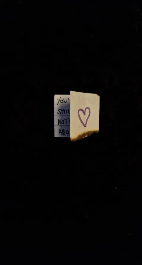The image shows a black background with two pieces of paper in the center. One is a small white piece of paper with a few lines of handwritten text partially visible. The other paper, also small and light-colored, has a purple drawn heart and a partially-burnt edge. The burnt edge has a dark color.