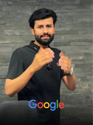 A man with dark hair and a beard stands in front of a textured gray wall. He is wearing a black polo shirt and a watch on his left wrist. The man is gesturing with both hands, and below him is the Google logo in the traditional colors.