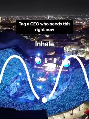An aerial view of a large outdoor venue at night, with a massive stage in the center and a sea of illuminated people filling the space. Overlaying this is a white, sinusoidal line. Above, a black banner contains text. Further text, 'Inhale,' is placed below the banner.