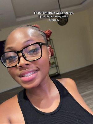 A selfie of a woman with dark skin, wearing black-rimmed glasses and a black tank top. She is smiling widely, with bright lipstick and subtle makeup. Her hair is styled in small braids with reddish-brown accents. The background is a softly lit room with neutral tones, featuring a glimpse of a television screen and a plant. Text overlay at the top of the image in white.