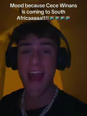 A selfie-style shot featuring a young man with dark hair and headphones, smiling with his mouth open. He is wearing a black shirt and a beaded necklace. The background has an orange-yellow tint, suggesting indoor lighting. Overlaid text says "Mood because Cece Winans is coming to South Africaaaaa!!!!" with South African flags.