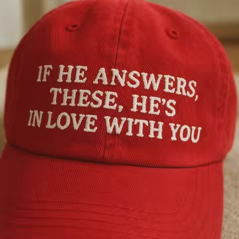 Close-up of a red baseball cap with white embroidered text that reads: "IF HE ANSWERS, THESE, HE'S IN LOVE WITH YOU". The cap is centered and fills most of the frame, with the curved brim partially visible. The background is slightly blurred, hinting at an indoor setting with neutral tones.