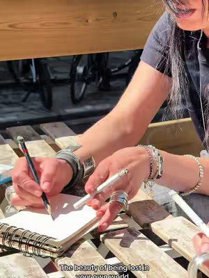 The cover image shows a person writing in a notebook with a pencil, likely outdoors. The person is wearing bracelets and rings; a second pencil is held by the other hand. Sunlight casts shadows on wooden slats. The text overlay says, 'The beauty of being lost in your own world.'