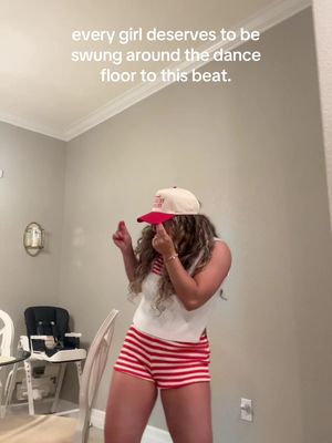 A woman is dancing in a room, wearing a baseball cap, striped shorts, and a tank top. Text overlayed on the image says "every girl deserves to be swung around the dance floor to this beat."
