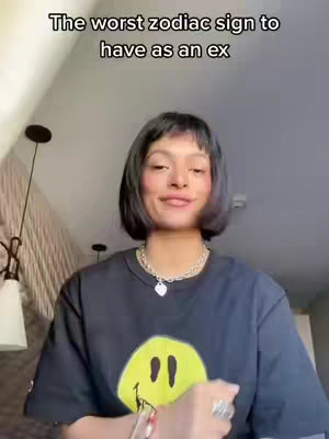 A woman with short black hair and a black smiley face t-shirt is centered in the frame. The background is a light-colored wall and ceiling with some visible architectural details. She is smiling slightly and wearing a necklace.