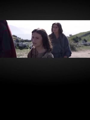 The image shows a scene from a film, likely depicting the young Jesus with a serious expression, accompanied by a woman in the background, possibly his mother. The setting appears to be outdoors, with a blurred background suggesting a natural environment. The color palette is muted, giving a slight historical feel.