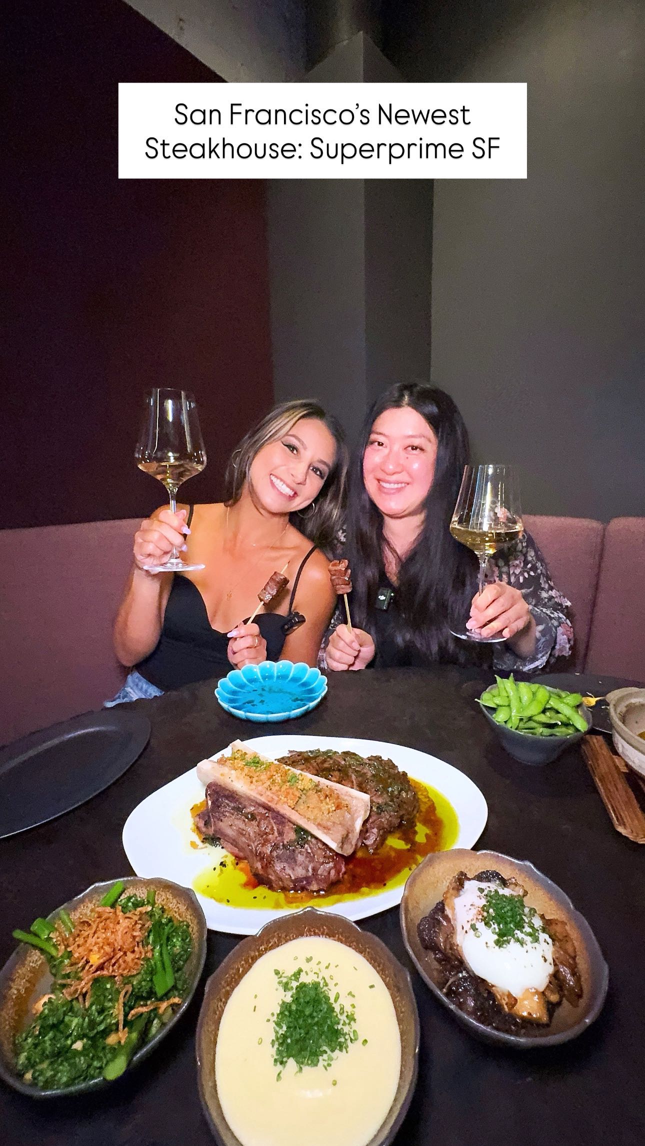 Two women are seated at a dark table in a restaurant; they are smiling at the camera while holding glasses of wine. Numerous dishes of food are on the table in front of them: a bone-in ribeye steak, several sides, plus plates of starters in the foreground. The background is a muted maroon.