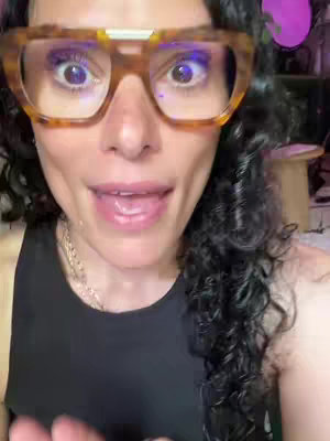A close-up shot of a woman with curly black hair, wearing large, tortoise-shell framed glasses with yellow accents and a black tank top. She is speaking, with her mouth slightly open and tongue visible. The background is out of focus, with hints of purple and other objects.