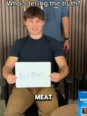 A young man sits in a chair, holding a whiteboard with the words "NUT Meat" written on it in marker. The word "MEAT" is also written in large bold letters at the bottom of the image. Behind him, another person stands, partially visible, with a navy shirt. The background consists of wooden paneling.