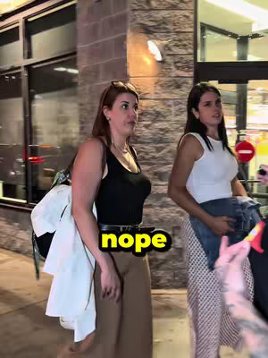 Two women are seen walking side-by-side in front of a building at what appears to be nighttime. The woman in the foreground is wearing a black top and brown pants, with sunglasses on her head. The image has a sticker overlay of 'nope', with a yellow background, positioned over her midsection. The second women is wearing a white tank top and a patterned skirt. The building has a sign in the background.