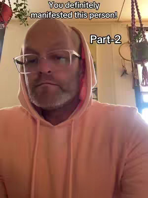 A medium shot features a man wearing glasses and a salmon-colored hoodie. The man's head and shoulders fills the frame. The text overlay reads "You definitely manifested this person!" and "Part-2". The background is softly blurred and includes visible decor, suggesting an indoor setting. The man has a beard and appears to be looking directly at the camera.
