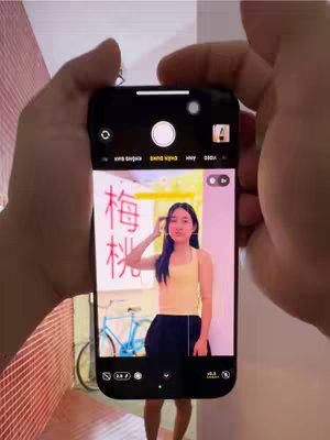 A person is holding a phone displaying a portrait photo of a woman. The phone screen shows a woman with long dark hair posing for a photo in what appears to be a cafe. The cafe has a colorful decor and a bicycle is visible behind the woman. The phone's camera interface shows a caption with Vietnamese text. The phone is held in two hands against a neutral background.