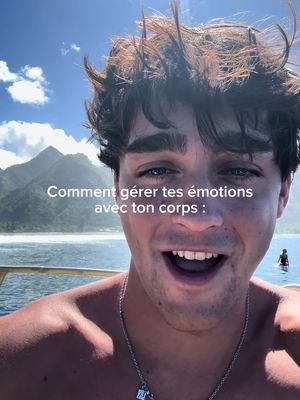 The cover image presents a cheerful, fair-skinned man, likely in his early twenties, against a scenic outdoor background. He has short, tousled brown hair and is smiling widely with his mouth open, revealing his teeth. The background features a bright blue sky, mountains in the mid-distance, and water in the foreground. The man is shirtless, and a silver necklace can be seen around his neck. Overlayed text in white font reads, 'Comment gérer tes émotions avec ton corps :'. The overall composition suggests a video related to emotional well-being in a natural setting.