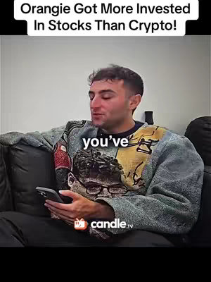 A man is seated on a black couch, looking towards the viewer while holding a phone. The background is a plain white wall. Text overlays appear on the image. The man wears an oversized, patterned sweater, and there is a logo in the lower corner.