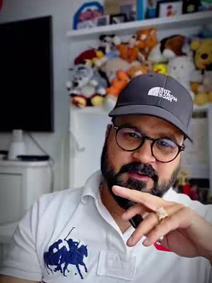 A man with glasses and a beard is centered in a casual, indoor shot. He wears a black cap with white text and a white polo shirt featuring a blue logo on the chest. A ring is on his right hand. The background, which includes soft toys and a TV, suggests a home setting.