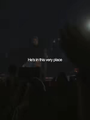 The image shows a dim, dark concert or event setting. Projected onto a surface, the text 'He's in this very place' is displayed. The background suggests a stage or performance area with figures in the dark. The overall lighting is focused and creates a sense of mystery and anticipation.
