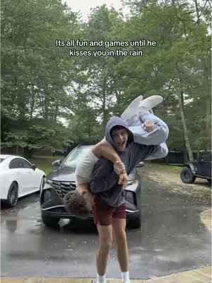 Two young men playfully engaged in a rain-soaked scene. One is carrying the other upside down over his shoulders, both appearing to be laughing. The background includes a car and lush trees, suggesting an outdoor environment.