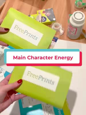 The image shows a close-up shot of hands holding two packages of FreePrints, a photo printing service. The packages are encased in bright green paper with the FreePrints logo in a white label. Some other items are visible in the background such as tape and glue.