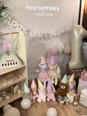 A staged scene celebrating a first birthday, featuring a collection of plush toys wearing party hats, scattered around a wooden shelf and the floor. A garland that says "Happy Birthday" hangs above the arrangement. Balloons and a large number one balloon float in the background.