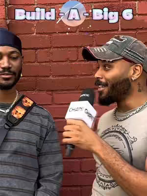 Two men are standing close together in front of a red brick wall. The man on the right is holding a microphone with a branded cover, while the man on the left is looking at the camera. The top of the image has the overlaid text 'Build-A-Big 6'.