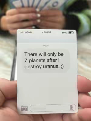 A hand holds a phone-shaped card displaying a text message conversation. The card appears to be a prop or a physical representation of a phone screen. The background is blurred, and the focus is on the hand and the card.