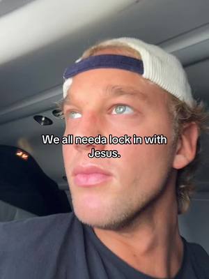 A close-up shot of a young man with fair skin, blue eyes, and blonde hair. He is wearing a white and navy baseball cap. The background is a blurred interior, possibly a plane cabin, with muted tones. Text overlayed on the image reads: 'We all need a lock in with Jesus.'