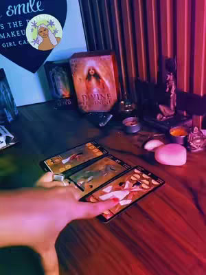 The image features a tarot reading setup. Three tarot cards are laid out, held by a person's hand, with the fingers and part of the wrist visible, the lighting is warm, with a table in focus. A wooden table is adorned with candles, a book, and what appears to be small, pink heart-shaped objects, adding to the mystical aesthetic.