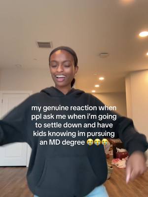 A young woman wearing a black hoodie is mid-motion in front of a light-colored wall and door frame. Text overlays the image conveying text. The composition is casual and candid, capturing a moment in a well-lit room.