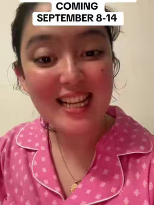 A selfie-style shot featuring a woman smiling and looking directly at the camera. She is wearing a pink, button-down shirt with a white collar and small, repeating white floral patterns. The background is a soft, off-white color. Text appears in a white box in the top middle.