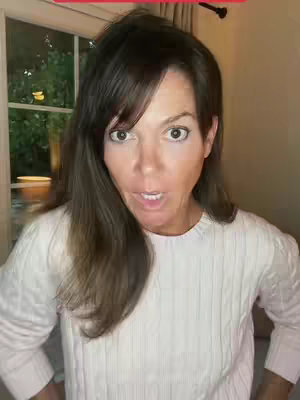 A woman with dark hair and wide eyes looks directly at the camera with an open mouth, as if she is surprised. She wears a white knit sweater. The background includes a window with greenery visible outside.
