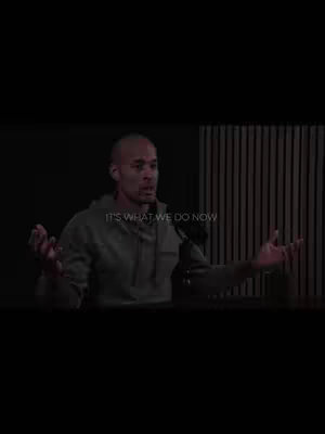 A man with a shaved head is centered in the frame, speaking into a microphone. The background consists of vertical wood panels. The text overlay says "IT'S WHAT WE DO NOW". The lighting is dim.