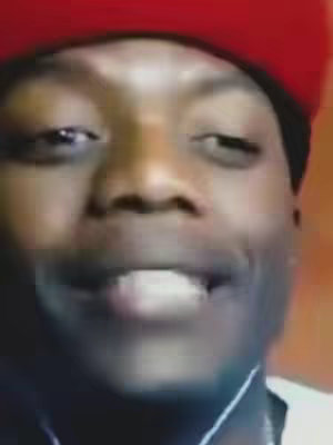 A close-up of a person's face from the chest up, shot in selfie mode. The person has dark skin and is wearing a red baseball cap. They are smiling, and their teeth are visible. The background is blurred and appears to be a warm tone, possibly orange or brown. The lighting seems to be natural, and the overall composition is simple, focusing entirely on the person's expression and face.