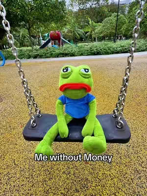 A plush frog character wearing a blue shirt sits on a playground swing. The background shows a park scene with trees, grass, and a slide.
