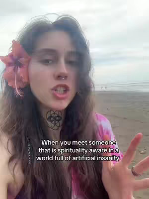 A young woman with long brown hair, a flower in her hair, and a tattoo on her neck is speaking near a beach. Overlayed text in white provides a caption about spiritual awareness.
