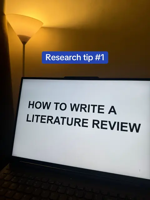 The image is a dimly lit indoor shot, likely in a room. The primary focus is a laptop displaying the text "HOW TO WRITE A LITERATURE REVIEW" in a large, clear, sans-serif font. The laptop is positioned at an angle, with only part of the keyboard visible. Above the laptop screen, in a blue rectangular box, reads the text "Research tip #1". In the background, a tall, cone-shaped lamp provides warm, diffused lighting, casting shadows on the wall. The overall composition is simple, emphasizing the laptop's screen and the instructional content displayed. The lighting suggests a nighttime or indoor setting, and the focus on the text implies a tutorial or educational context.