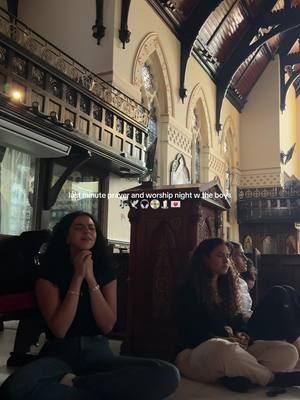 The image captures a dimly lit interior of a church. The main focus is on several young women seated on the floor, their hands clasped in prayer, indicating a religious or worship gathering. The backdrop includes high ceilings, arched windows, and ornate architectural details, creating a solemn ambiance. The overall tone is intimate and serene.