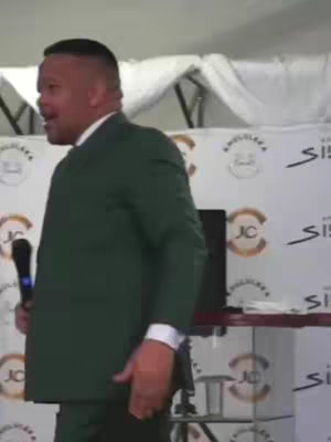 A man in a dark green suit is speaking into a microphone. He is on a stage with a white backdrop that has logos printed on it. The lighting appears to be bright.