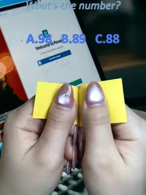 A person's hands, with long, glossy pink nails, holding a small yellow notepad open, with a screen in the background showing a website for the website Pexel with blue options.