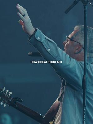 A medium shot captures a man with glasses singing and raising his arm. The lighting is low, casting shadows and emphasizing the subject's face and hand. The text overlay at the bottom of the image reads 'HOW GREAT THOU ART.' This suggests a religious or worship theme, likely from a church service or similar event. The overall composition is evocative of a live performance.