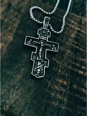 Close-up shot of an Orthodox cross necklace resting on a dark, textured surface. The cross is silver-colored with intricate details, including a figure of Jesus. The surface appears to be wooden or stone, with a dark, grainy texture. The necklace chain is also visible.
