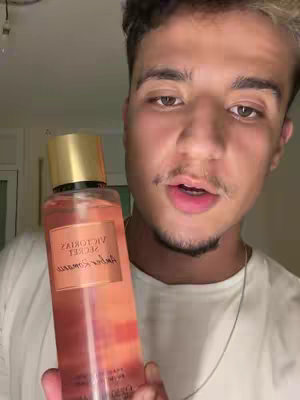 A man is holding a bottle of Victoria's Secret Amber Romance body mist in his left hand, presenting toward the camera. The bottle is filled with a gradient of orange and pink, and the label is cream with black text. The man's face is close to the camera, with brown eyes, a small mustache, and a goatee. He has short, dark hair, and a pale complexion. He is wearing a white t-shirt and a silver chain, and is inside, possibly lit by room light.
