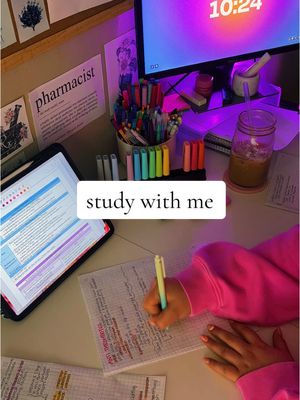 A well-lit, top-down shot of a study space. On a white desk are school supplies and a tablet displaying notes. A person is writing with a light green pen on a piece of paper. In the background, a computer shows a clock, study notes, and a mug of coffee is alongside a pen holder full of vibrant colored pens.