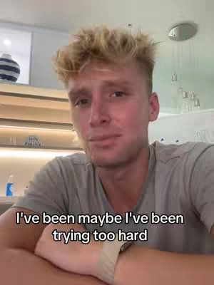 A selfie-style shot featuring a young man. He has light-colored hair and is positioned with his upper body visible. The background is a kitchen setting with hints of a microwave and decorative items. Overlay text appears at the bottom, with the man looking at the camera with a slight smile, wearing a grey t-shirt.