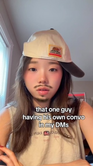 A young woman with long hair is wearing a baseball cap and a sleeveless top. She has applied a fake mustache and goatee. The text overlay reads: "that one guy having his own convo in my DMs".