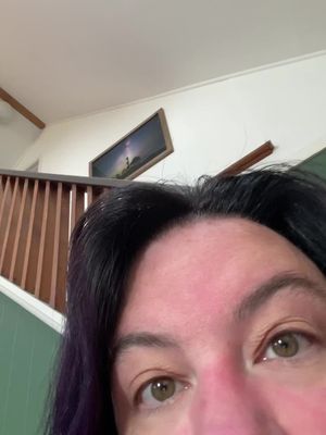 A selfie from the chest up, capturing a person's eyes and the space above their forehead. The person has dark hair and is looking towards the camera at an angle, with a glimpse of a staircase and a framed artwork in the background.