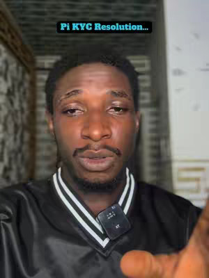 A close-up shot of a man with dark skin and a serious expression. He has a trimmed mustache and goatee and is wearing a black shirt with a white-lined collar. A small microphone is clipped to his shirt. Above his head, a text overlay reads "Pi KYC Resolution..."