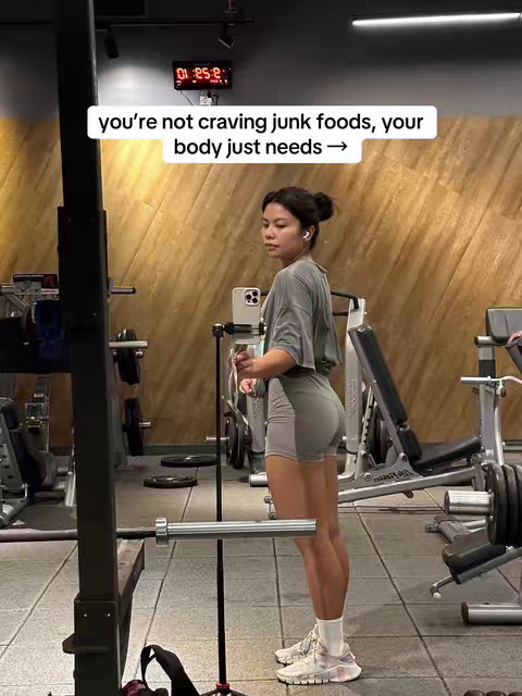 A woman in workout attire stands sideways in a gym setting. She is wearing a grey t-shirt and shorts. The background features exercise equipment and wooden wall panels.