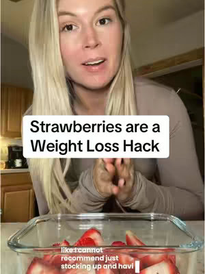 A woman with blonde hair is filming a video in what appears to be a kitchen. Text overlay 'Strawberries are a Weight Loss Hack' is prominently displayed across her upper chest. A glass container filled with sliced strawberries is visible below the text and the woman. Other kitchen elements are in the blurred background.