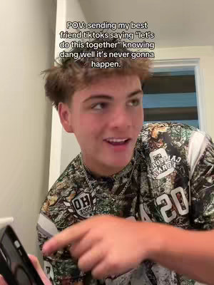 A young man with short brown hair is holding a phone with the screen visible, with a light colored wall in front of him, along with a window visible behind him. The man is wearing a camo print shirt and is smiling.